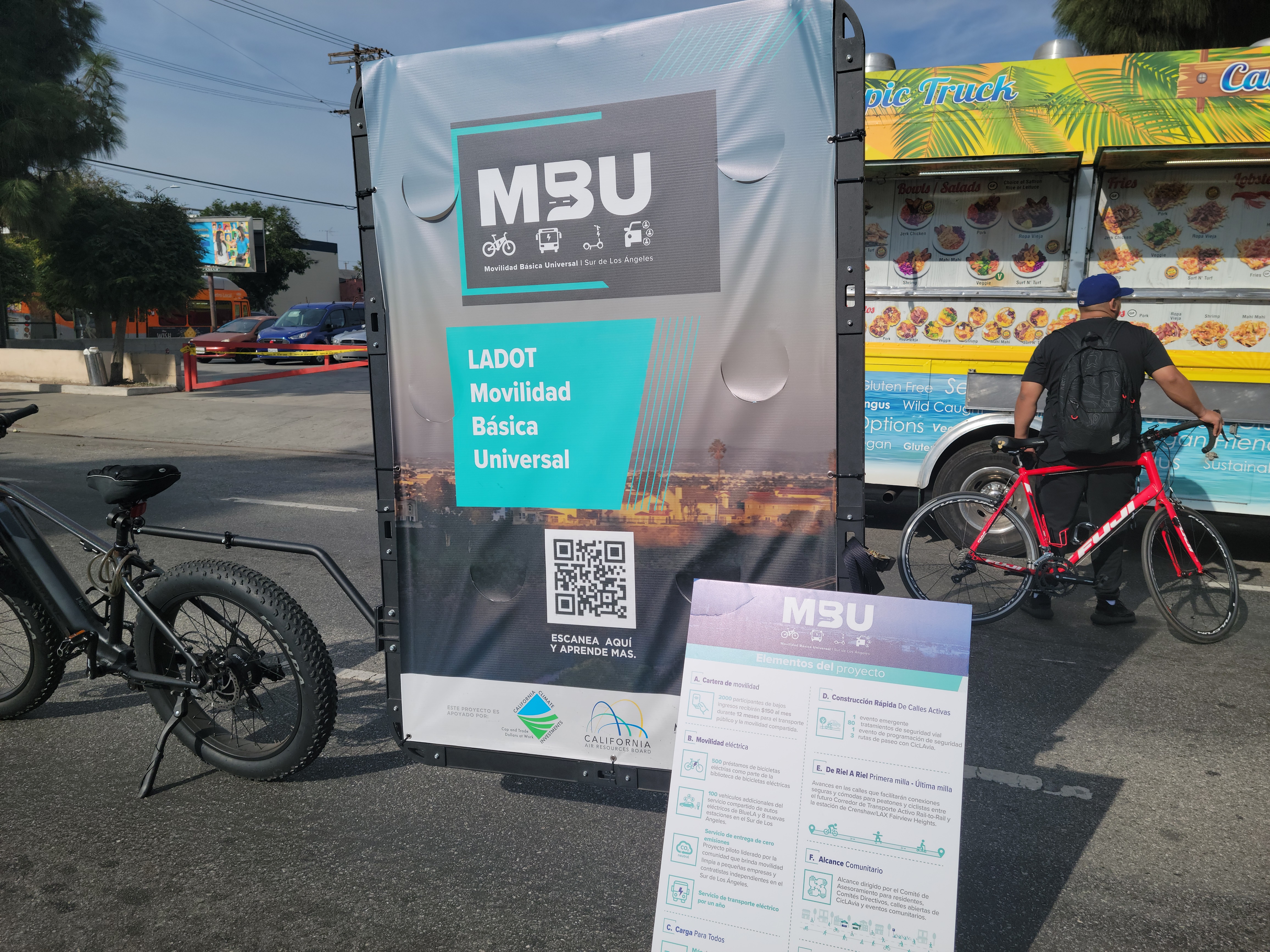 Black E bike pulling a mobile banner with Mobilidad Basico Universal in front of a food truck and a man on a red bicycle